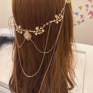 May include: A gold-colored headpiece with a floral design and dangling chains. The headpiece features a central teardrop-shaped jewel and delicate floral accents along the band. The chains hang down, adding movement and elegance to the hair accessory.