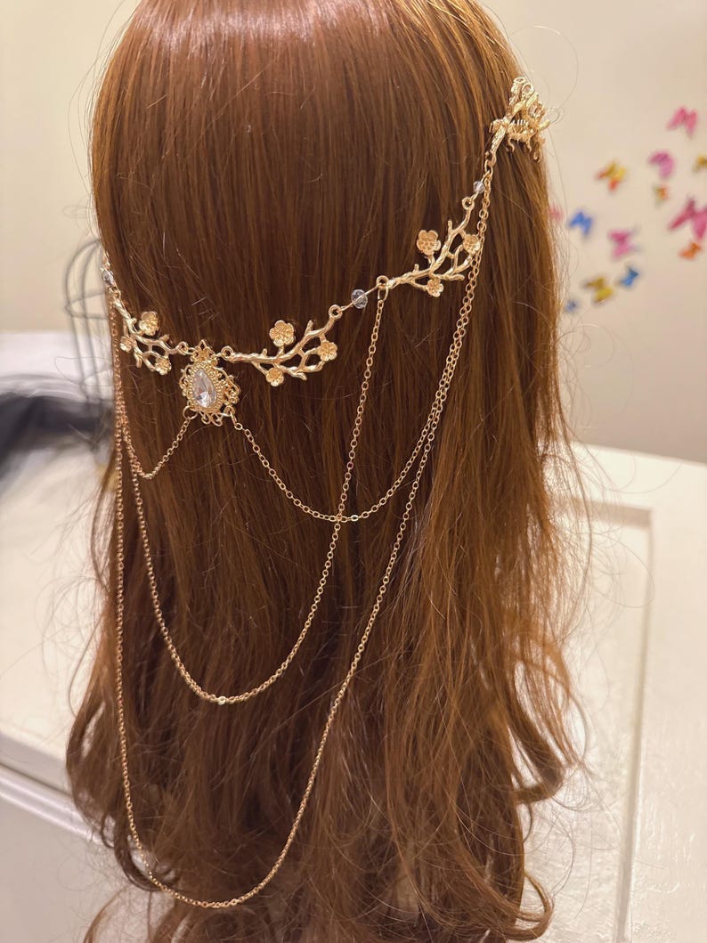 May include: A gold-colored headpiece with a floral design and dangling chains. The headpiece features a central teardrop-shaped jewel and delicate floral accents along the band. The chains hang down, adding movement and elegance to the hair accessory.