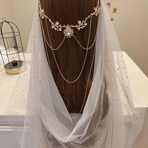 May include: A gold-colored headpiece with floral and chain details, featuring a central teardrop-shaped gem. A sheer white veil with silver glitter dots drapes over the head. The headpiece is designed for special occasions.