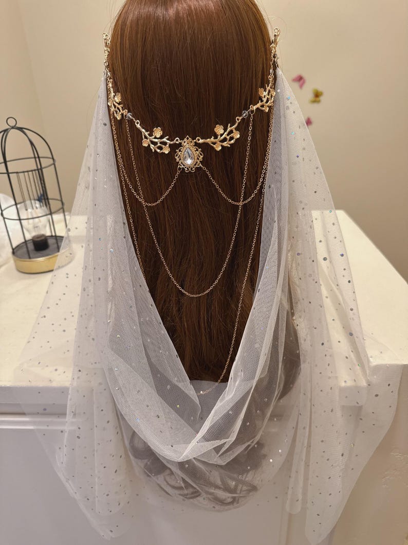 May include: A gold-colored headpiece with floral and chain details, featuring a central teardrop-shaped gem. A sheer white veil with silver glitter dots drapes over the head. The headpiece is designed for special occasions.