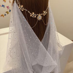 May include: A gold-toned headpiece with floral and chain details, adorned with a central teardrop-shaped gem. A sheer, white veil with scattered iridescent sequins drapes down, creating a delicate, ethereal look.