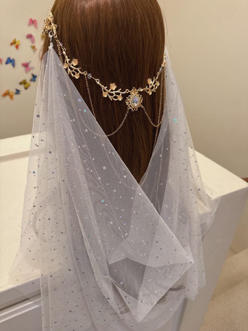 May include: A gold-toned headpiece with floral and chain details, adorned with a central teardrop-shaped gem. A sheer, white veil with scattered iridescent sequins drapes down, creating a delicate, ethereal look.