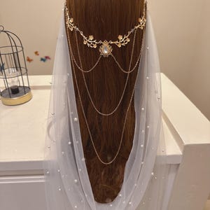 May include: A gold-colored bridal headpiece with a floral design and dangling chains, paired with a white veil adorned with pearls. The headpiece features a central jewel and is worn over brown hair.