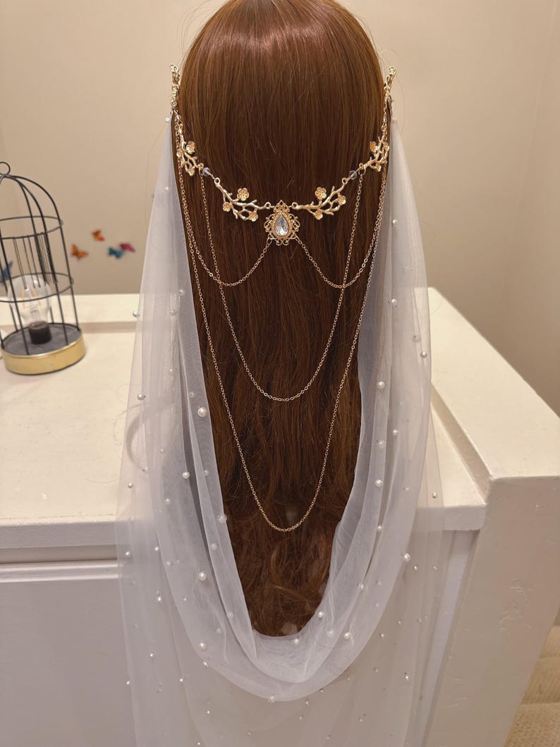 May include: A gold-colored bridal headpiece with a floral design and dangling chains, paired with a white veil adorned with pearls. The headpiece features a central jewel and is worn over brown hair.