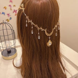 May include: A gold-colored headpiece with a celestial theme. The headpiece features a crescent moon, dangling teardrop-shaped crystals, and leaf accents. The headpiece is designed to be worn on the head and is likely for special occasions.