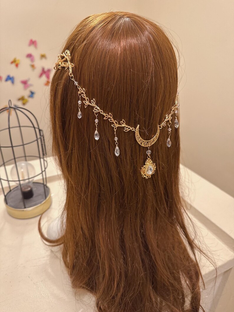 May include: A gold-colored headpiece with a celestial theme. The headpiece features a crescent moon, dangling teardrop-shaped crystals, and leaf accents. The headpiece is designed to be worn on the head and is likely for special occasions.