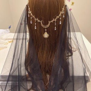May include: A gold-toned headpiece with a crescent moon and teardrop accents, paired with a sheer black veil. The headpiece features a delicate floral design and a central pendant. The veil drapes down the back, creating an ethereal look.