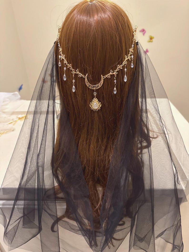May include: A gold-toned headpiece with a crescent moon and teardrop accents, paired with a sheer black veil. The headpiece features a delicate floral design and a central pendant. The veil drapes down the back, creating an ethereal look.