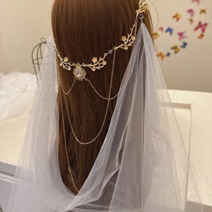 May include: A gold-colored bridal headpiece with a floral design and dangling chains. The headpiece features a central jewel and is paired with a sheer white veil. The headpiece is worn on a mannequin with brown hair.