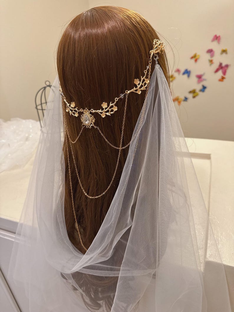 May include: A gold-colored bridal headpiece with a floral design and dangling chains. The headpiece features a central jewel and is paired with a sheer white veil. The headpiece is worn on a mannequin with brown hair.
