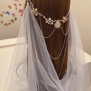 May include: A gold-colored bridal headpiece with a floral design and a central teardrop-shaped gem. Delicate chains hang from the headpiece, and a sheer white veil drapes down. The headpiece is worn on brown hair.