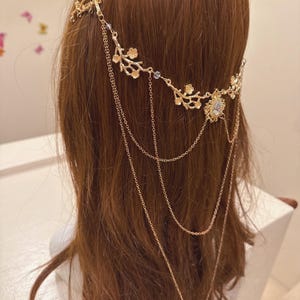 May include: Gold-toned headpiece with a floral design and dangling chains. The headpiece features delicate branches with small flowers and a central jewel. The chains cascade down, adding an elegant touch to the hair accessory.