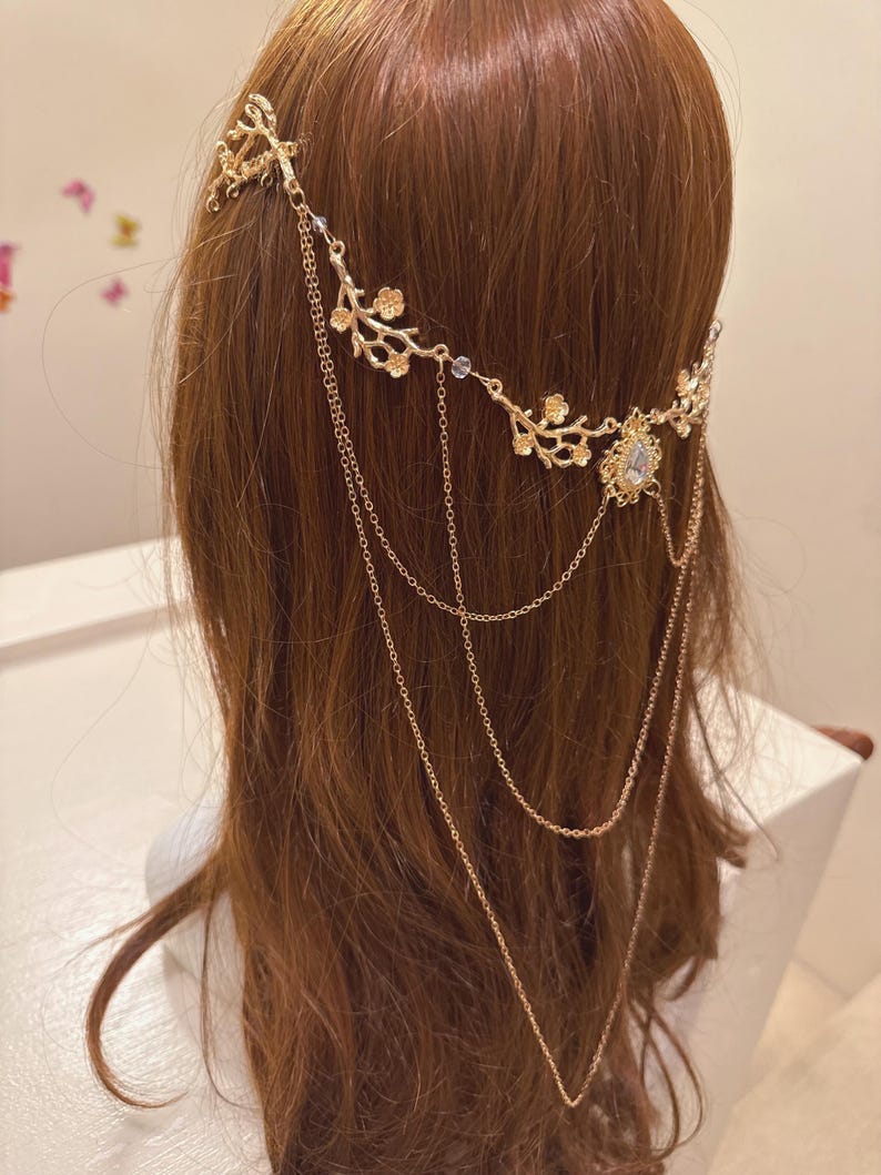 May include: Gold-toned headpiece with a floral design and dangling chains. The headpiece features delicate branches with small flowers and a central jewel. The chains cascade down, adding an elegant touch to the hair accessory.
