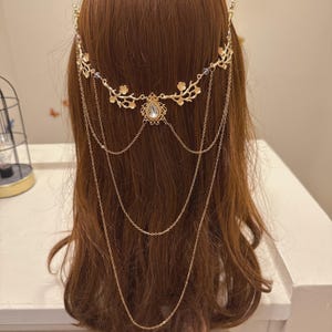May include: A gold-colored headpiece with a floral design and dangling chains. The headpiece features a central teardrop-shaped gem and is worn on brown hair. The design includes delicate floral accents and multiple chains.