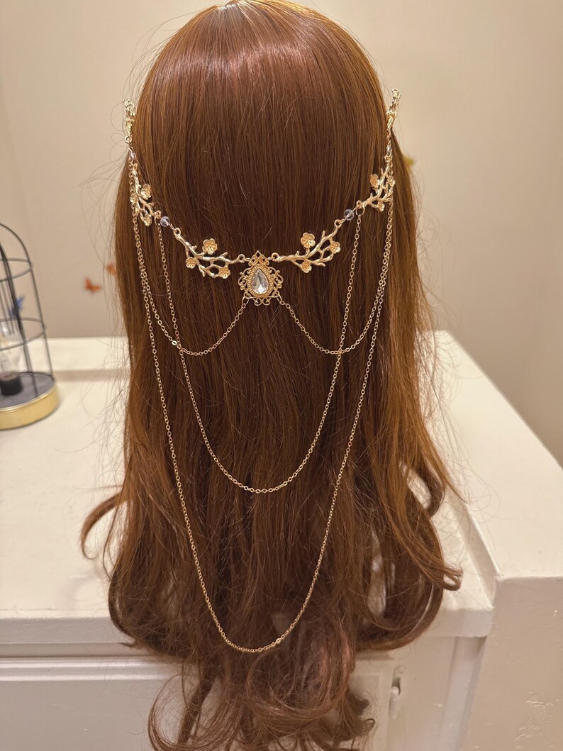 May include: A gold-colored headpiece with a floral design and dangling chains. The headpiece features a central teardrop-shaped gem and is worn on brown hair. The design includes delicate floral accents and multiple chains.