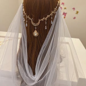 May include: A gold-colored headpiece with a crescent moon and teardrop-shaped crystal pendants. The headpiece is adorned with delicate leaf designs and is paired with a sheer white veil, creating an ethereal look. The headpiece is designed to be worn on the head.