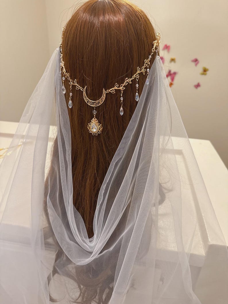 May include: A gold-colored headpiece with a crescent moon and teardrop-shaped crystal pendants. The headpiece is adorned with delicate leaf designs and is paired with a sheer white veil, creating an ethereal look. The headpiece is designed to be worn on the head.