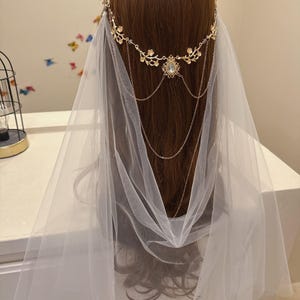 May include: A bridal headpiece featuring a gold-toned floral crown with a central jewel and delicate chain accents. A sheer white veil cascades down, creating a soft, ethereal look. The headpiece is designed for weddings and special occasions.