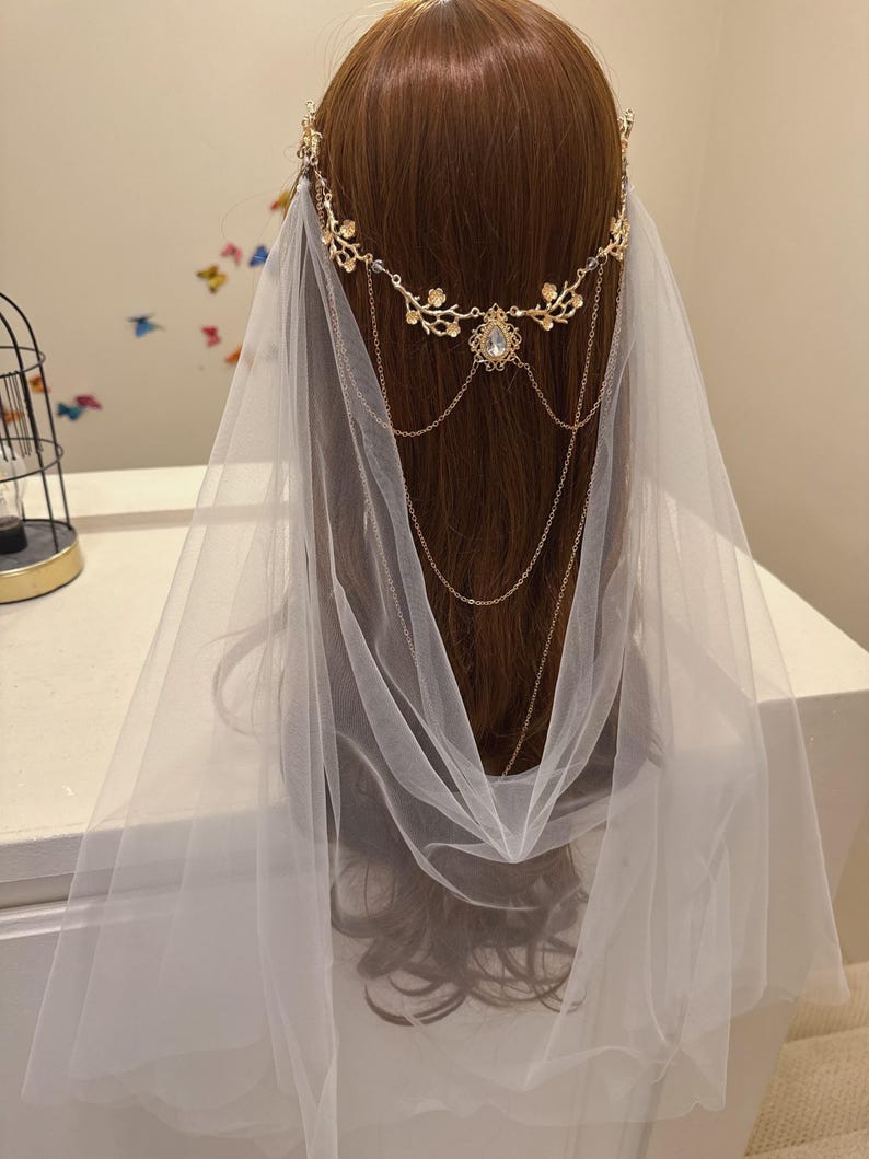May include: A bridal headpiece featuring a gold-toned floral crown with a central jewel and delicate chain accents. A sheer white veil cascades down, creating a soft, ethereal look. The headpiece is designed for weddings and special occasions.