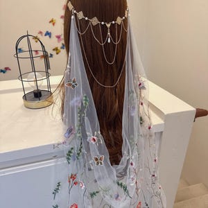 May include: A gold-toned headpiece with a delicate veil. The headpiece features square and pearl accents, with cascading chains. The sheer veil is adorned with colorful embroidered flowers and butterflies, creating an ethereal look.