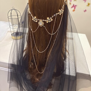 May include: A gold-colored headpiece with floral and chain details, featuring a central crystal pendant. A black tulle veil drapes down, creating a layered effect. The headpiece is worn on brown hair.