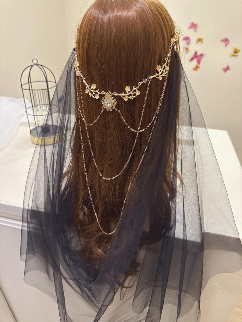 May include: A gold-colored headpiece with floral and chain details, featuring a central crystal pendant. A black tulle veil drapes down, creating a layered effect. The headpiece is worn on brown hair.