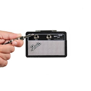 May include: A black and silver Fender Mini Twin Amp with two input jacks and a speaker grill. The amp is small and compact, with the text "Mini Twin Amp" and "Fender Musical Instruments" printed on the front. A hand holds a guitar cable.