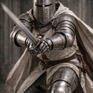 May include: A medieval knight in full armor, holding a long sword. The knight's helmet, shoulder plates, and leg guards are made of metal. A white cape and tunic with a red cross are visible. The image includes the text "Medieval Store".