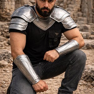 May include: A person wearing silver metal armor, including shoulder pauldrons and arm bracers, over a black t-shirt and jeans. The image includes the text "MEDIEVAL STORE" in a shield logo. The setting is outdoors with stone ruins.