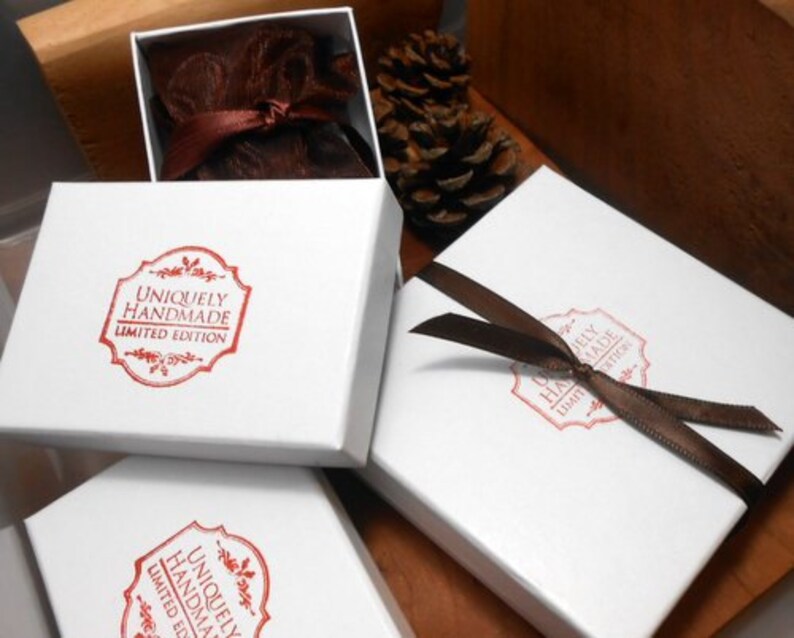 以下が含まれることがあります： 「Uniquely Handmade Limited Edition」と書かれた赤いスタンプが押された白い正方形の箱。1つの箱は開いており、茶色のリボンで結ばれた茶色の布製のポーチが見えます。別の箱には茶色のリボンが巻かれています。木製の表面にあります。