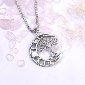 May include: A silver necklace with a crescent moon pendant, featuring a tree of life design and iridescent details. The chain is delicate. The background shows pink gemstones and a soft, feathery element. The necklace is a piece of jewellery.