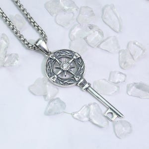 Puede incluir: Un collar de tono plateado con un colgante en forma de llave. La cabeza de la llave es un diseño de brújula circular con los puntos cardinales N, S, E y O, una piedra clara central y detalles de nudos intrincados. La cadena es de eslabones cuadrados.