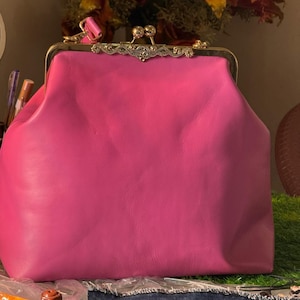 Könnte beinhalten: Eine leuchtend pinkfarbene Handtasche mit einem goldfarbenen Verschluss. Der Verschluss weist aufwendige Details und einen Kiss-Lock-Verschluss auf. Die Tasche hat eine abgerundete Form und scheint aus Leder oder einem ähnlichen Material zu bestehen. Im Hintergrund ist ein Blumenarrangement zu sehen.