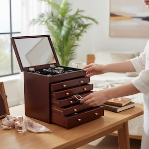 May include: A dark brown wooden jewelry box with a mirrored lid and multiple drawers. The drawers are open, revealing jewelry inside. The box sits on a wooden table, with a person's hands opening a drawer. Gold-colored knobs adorn the drawers.