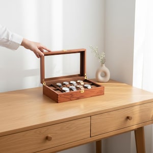 May include: A wooden watch display case with a clear glass top, showcasing several wristwatches. The case is open, revealing the watches nestled in individual compartments. The wood has a warm, reddish-brown tone, and the case is set on a light-colored wooden desk.