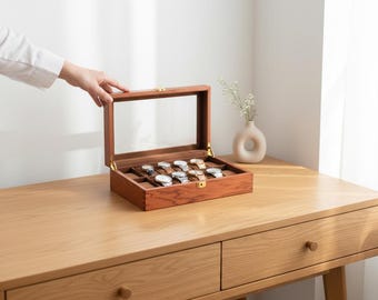 May include: A wooden watch display case with a clear glass top, showcasing several wristwatches. The case is open, revealing the watches nestled in individual compartments. The wood has a warm, reddish-brown tone, and the case is set on a light-colored wooden desk.