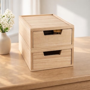 May include: A two-drawer wooden storage unit with a light oak finish. The drawers have recessed handles and are stacked on top of each other. The unit is sitting on a wooden surface, with a vase of white flowers in the background.