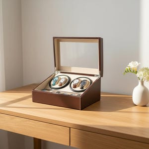 May include: A brown leather watch winder with a clear glass lid, holding four watches. The interior has a beige lining and wooden watch holders. The winder sits on a wooden desk, with a small vase of flowers to the right.