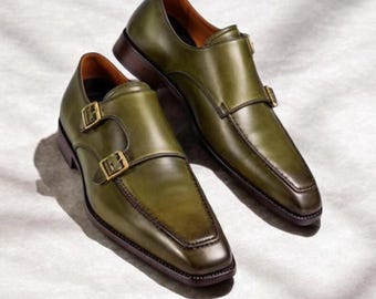 Könnte beinhalten: Ein Paar olivgrüne Lederschuhe mit Doppel-Monk-Riemen, quadratischer Zehenpartie, dunkelbrauner Sohle und zwei goldfarbenen Metallschnallen an jedem Schuh. Das Leder hat eine glatte Oberfläche mit subtilen, tonalen Schattierungen.