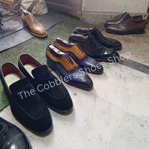 Pu&ograve; includere: Una collezione di scarpe da uomo, tra cui mocassini e scarpe eleganti, in nero, marrone e blu. Il testo "The Cobblers Shoes Shop" &egrave; visibile su alcune scarpe. Le scarpe sono disposte su un pavimento piastrellato.