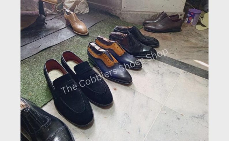 Pu&ograve; includere: Una collezione di scarpe da uomo, tra cui mocassini e scarpe eleganti, in nero, marrone e blu. Il testo "The Cobblers Shoes Shop" &egrave; visibile su alcune scarpe. Le scarpe sono disposte su un pavimento piastrellato.