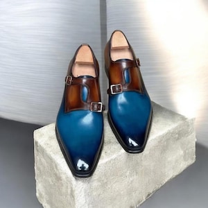Pu&ograve; includere: Un paio di scarpe eleganti in pelle bicolore. Le scarpe presentano una punta blu che sfuma verso il nero, con un cinturino in pelle marrone e fibbia. Sono esposte su una superficie di pietra grigia.