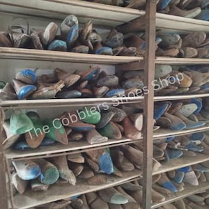 Pu&ograve; includere: Una scaffalatura in legno piena di forme per scarpe. Le forme sono di varie dimensioni e colori, tra cui blu, verde e marrone. Il testo "The Cobblers Shoes Shop" &egrave; visibile. Gli scaffali sono in legno e il telaio in metallo.