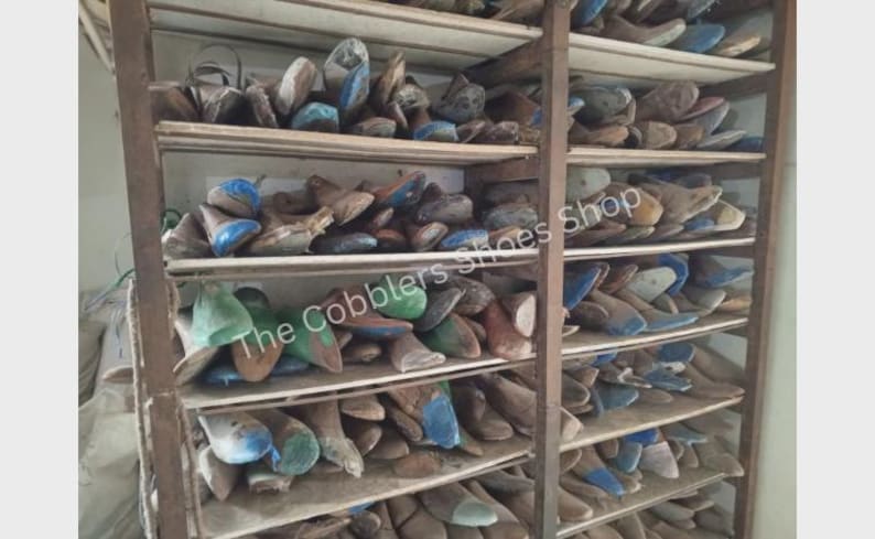 Pu&ograve; includere: Una scaffalatura in legno piena di forme per scarpe. Le forme sono di varie dimensioni e colori, tra cui blu, verde e marrone. Il testo "The Cobblers Shoes Shop" &egrave; visibile. Gli scaffali sono in legno e il telaio in metallo.
