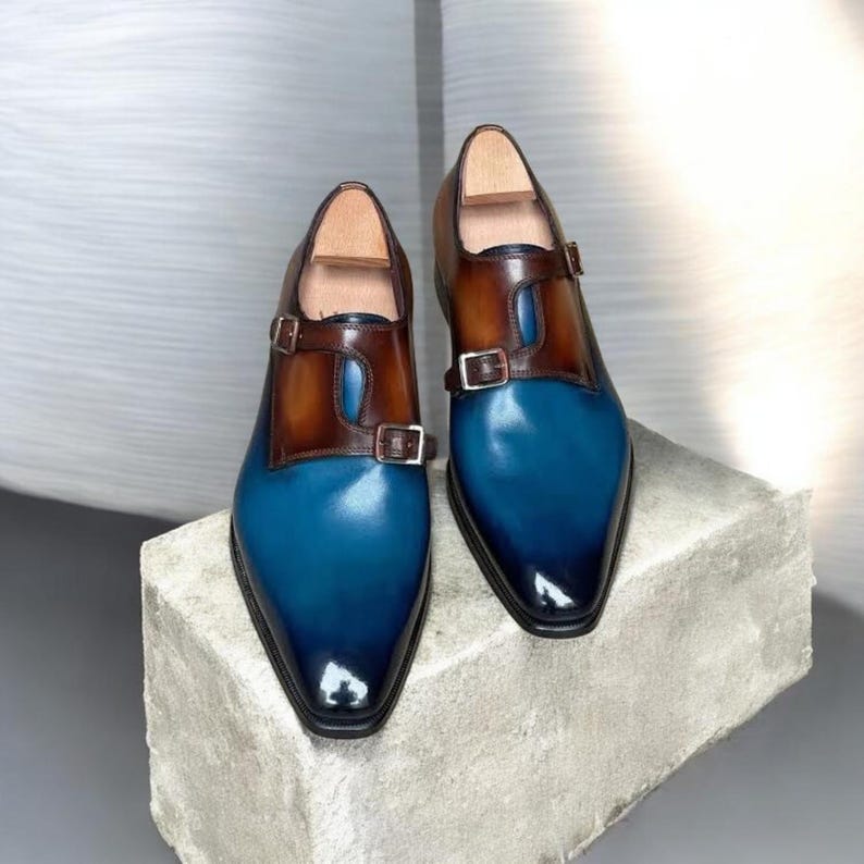 Pu&ograve; includere: Un paio di scarpe eleganti in pelle bicolore. Le scarpe presentano una punta blu che sfuma verso il nero, con un cinturino in pelle marrone e fibbia. Sono esposte su una superficie di pietra grigia.