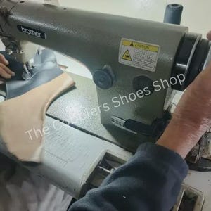 Pu&ograve; includere: Primo piano di una macchina da cucire industriale Brother in uso. La macchina &egrave; grigia e sta cucendo pezzi di pelle e tessuto. Il testo "The Cobblers Shoes Shop" &egrave; visibile.