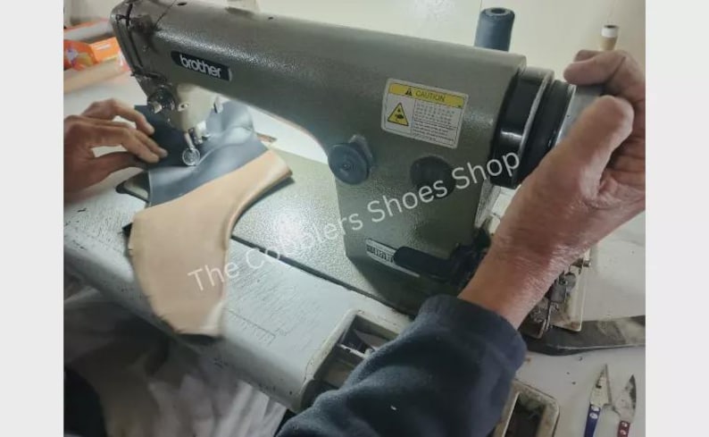 Pu&ograve; includere: Primo piano di una macchina da cucire industriale Brother in uso. La macchina &egrave; grigia e sta cucendo pezzi di pelle e tessuto. Il testo "The Cobblers Shoes Shop" &egrave; visibile.