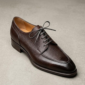 Puede incluir: Un zapato de vestir de cuero marrón con una superficie texturizada y cordones grises oscuros. El zapato presenta un diseño clásico con punta afilada y tacón bajo, adecuado para ocasiones formales. Mide aproximadamente 30 cm de largo.
