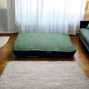 May include: A large, rectangular floor cushion with a knitted green top and a black base. The cushion sits on a hardwood floor, with a fluffy, light-colored rug in front of it. Sheer white curtains are in the background.