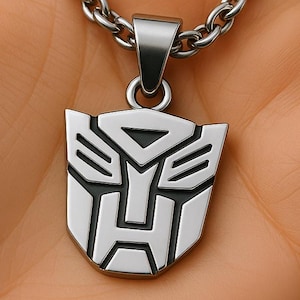 May include: A silver pendant necklace with the Autobot symbol. The pendant, in a shield shape with black and silver detailing, hangs from a chain. The necklace is shown against a neutral background.